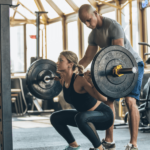 Woman peforming strength training high intensity deep barbell squats in gym with personal trainer standing behind spotting to ensure safety and technique