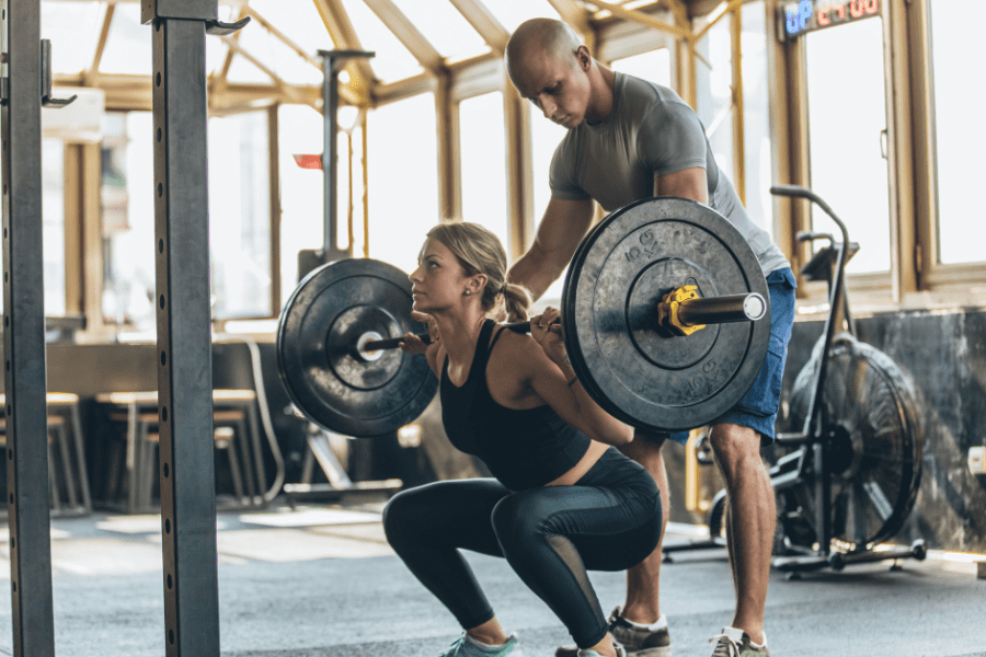 Woman peforming strength training high intensity deep barbell squats in gym with personal trainer standing behind spotting to ensure safety and technique